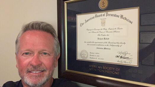 Dr. Kellett Man poses in front of a framed certificate from the American Board of Preventive Medicine on a wall.
